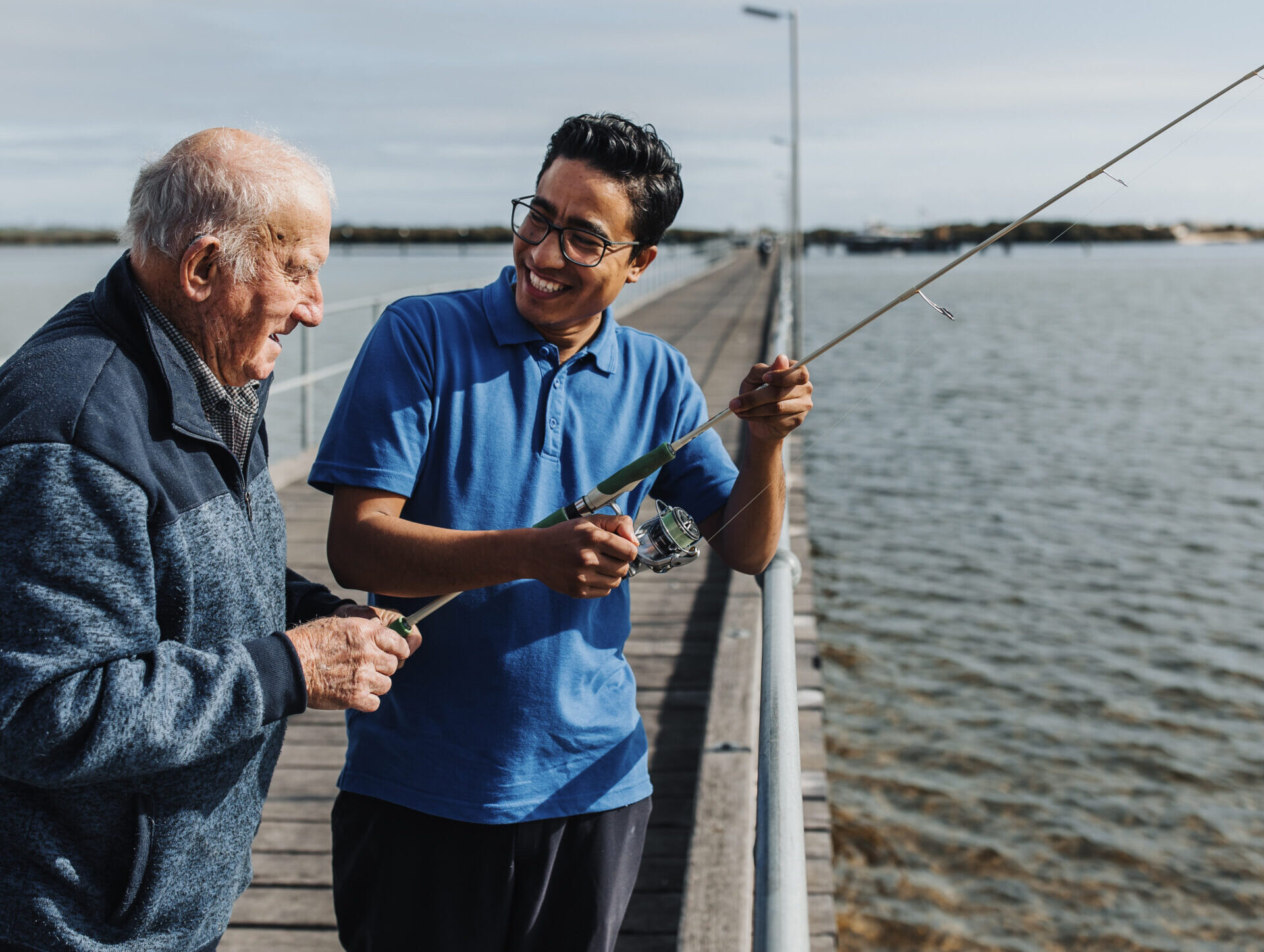 Older man and younger companion smiling while fishing together on a jetty.